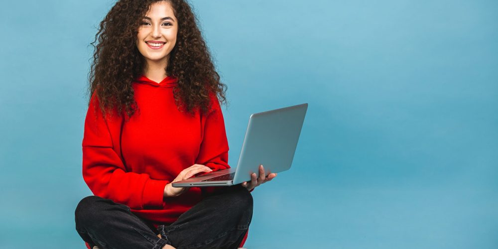 Happy young curly beautiful woman sitting on the floor with crossed legs and using laptop isolated on blue background.